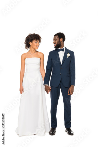 cheerful african american bride and bridegroom holding hands while standing isolated on white