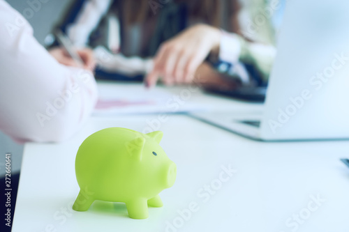 Two businesswomen discuss business graphics in the background, green piggy Bank in focus in the foreground.