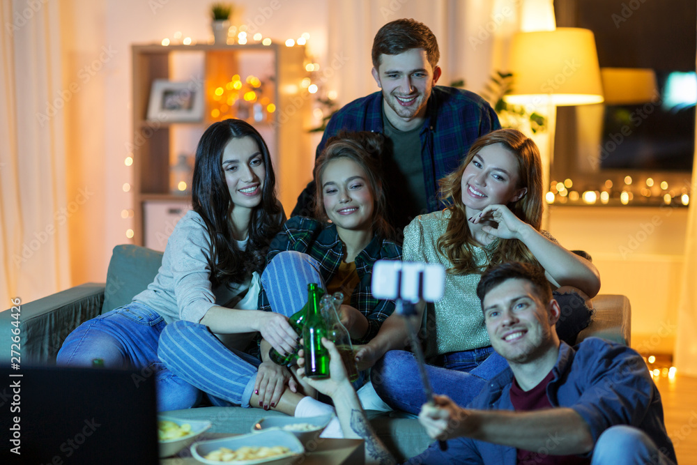 friendship, people, technology and entertainment concept - happy friends with snacks and non-alcoholic drinks watching tv and taking picture by selfie stick at home in evening
