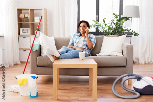 household and technology concept - happy african american woman or housewife resting after home cleaning and calling on smartphone photo