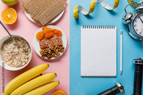 top view of fresh fruits, crispbread and breakfast cereal on pink and blank notebook, dumbbells and measuring tape on blue background