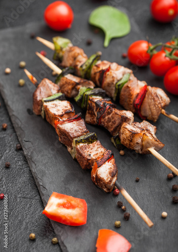Grilled pork and chicken kebab with paprika on stone chopping board with salt, pepper and tomatoes on black background.