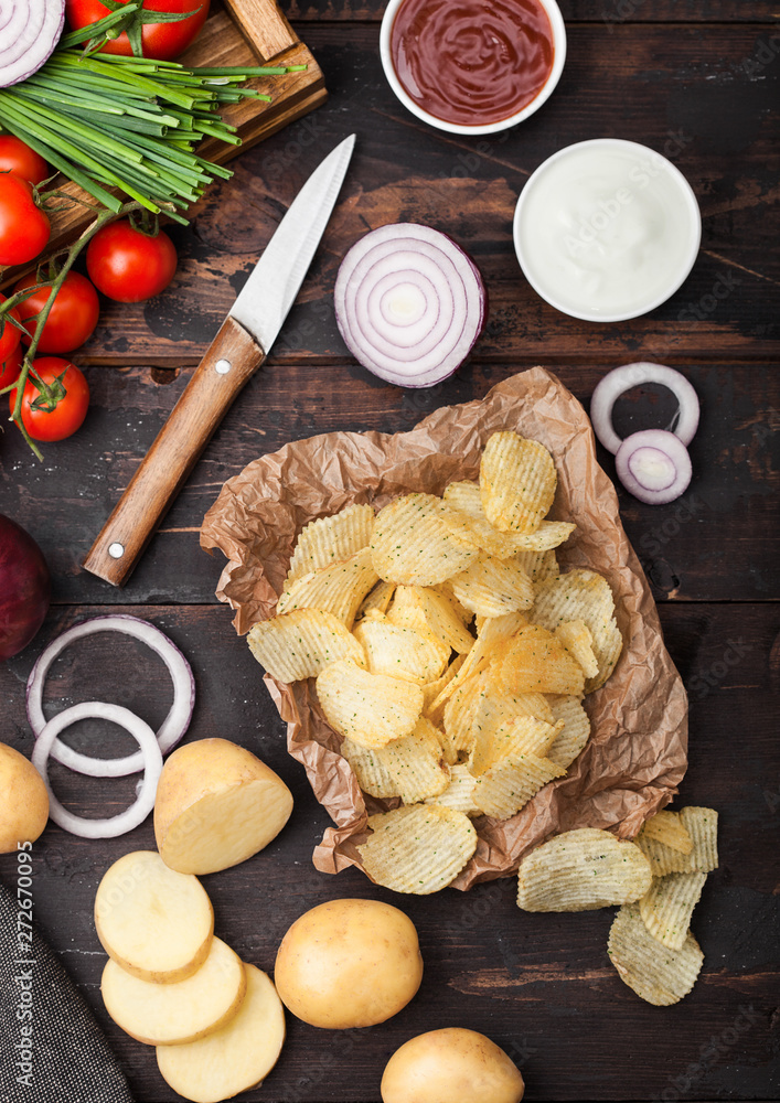 Fresh organic homemade potato crisps chips with sour cream and red onions on paper on dark wooden background. Tomatoes with green onion and bowl of ketchup.