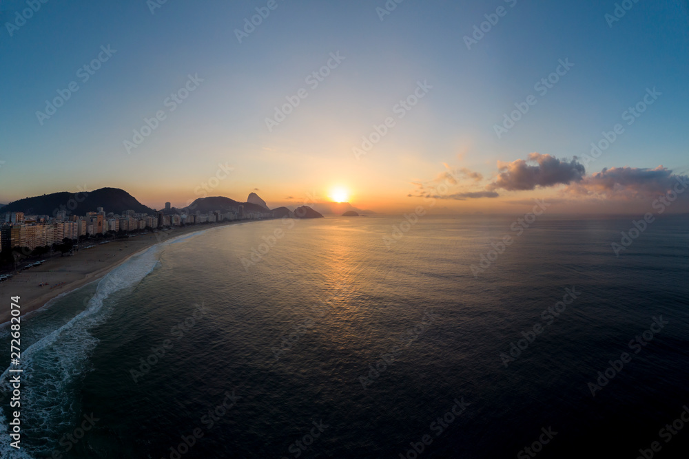 Aerial view of Copacabana beach and neighbourhood at early sunrise with the sun on the horizon next to the Sugarloaf mountain against a colourful blue sky seen from the ocean