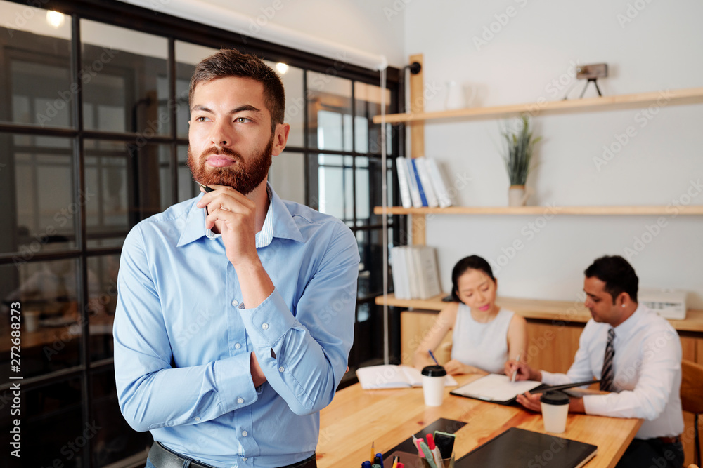 Young bearded businessman standing at office and thinking over something with his partners sitting at the table and working together in the background Young bearded businessman standing at office and thinking over something with his partners sitting at the table and working together in the background