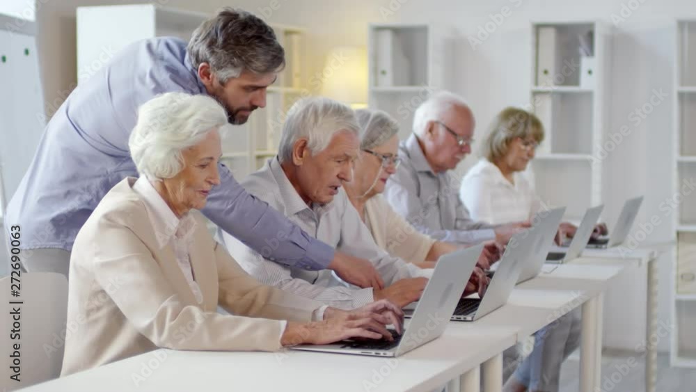 Medium shot of bearded middle-aged male teacher helping elderly people sitting at desks and typing on laptops in technology class Medium shot of bearded middle-aged male teacher helping elderly people sitting at desks and typing on laptops in technology class