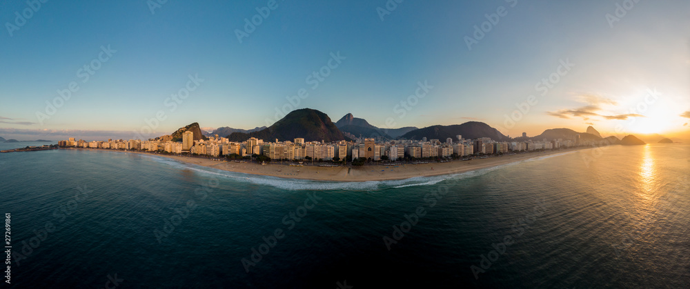 Curved full aerial view of Copacabana beach and neighbourhood at early sunrise flanked by the Sugarloaf mountain on the right and Copacabana fort on the left against a blue sky seen from the ocean