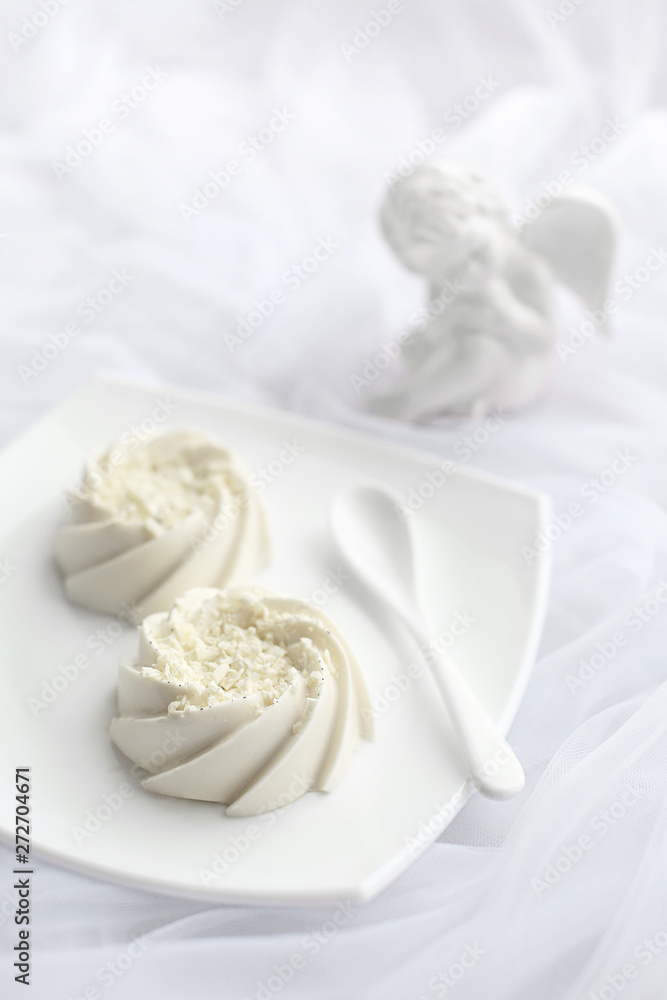 Light airy dessert panna cotta mousse with white chocolate and vanilla on top, on white ceramic plate and ceramic spoon and porcelain angel statuette. Monochrome Light airy dessert panna cotta mousse with white chocolate and vanilla on top, on white ceramic plate and ceramic spoon and porcelain angel statuette. Monochrome