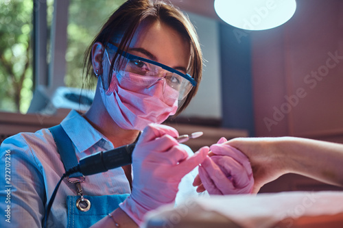 Young cheerful manicurist in glasses and protective mask is working at her workplace with customer.