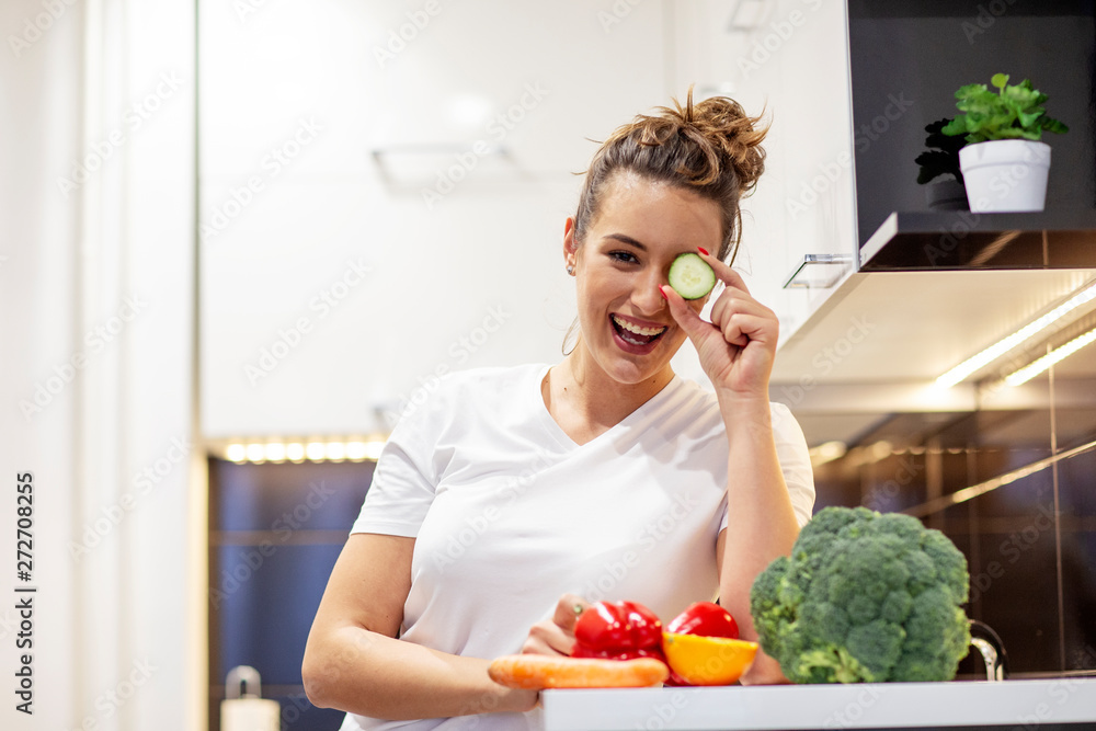 Beautiful girl in kitchen with cucumber in front of her eye having ful while prepare vegan meal