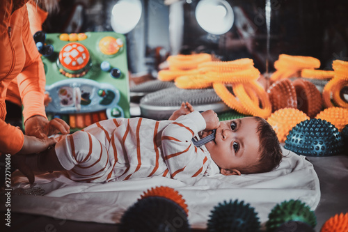 Happy smiling cute toddler is lying on the special table surrounded by ortopedic toys while expirienced masseur making massage for him. photo