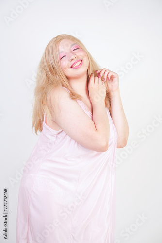 Charming plus size blonde asian girl in pink pretty silk nightgown standing and happy posing on white background in Studio alone. Chubby attractive woman in cute dress.