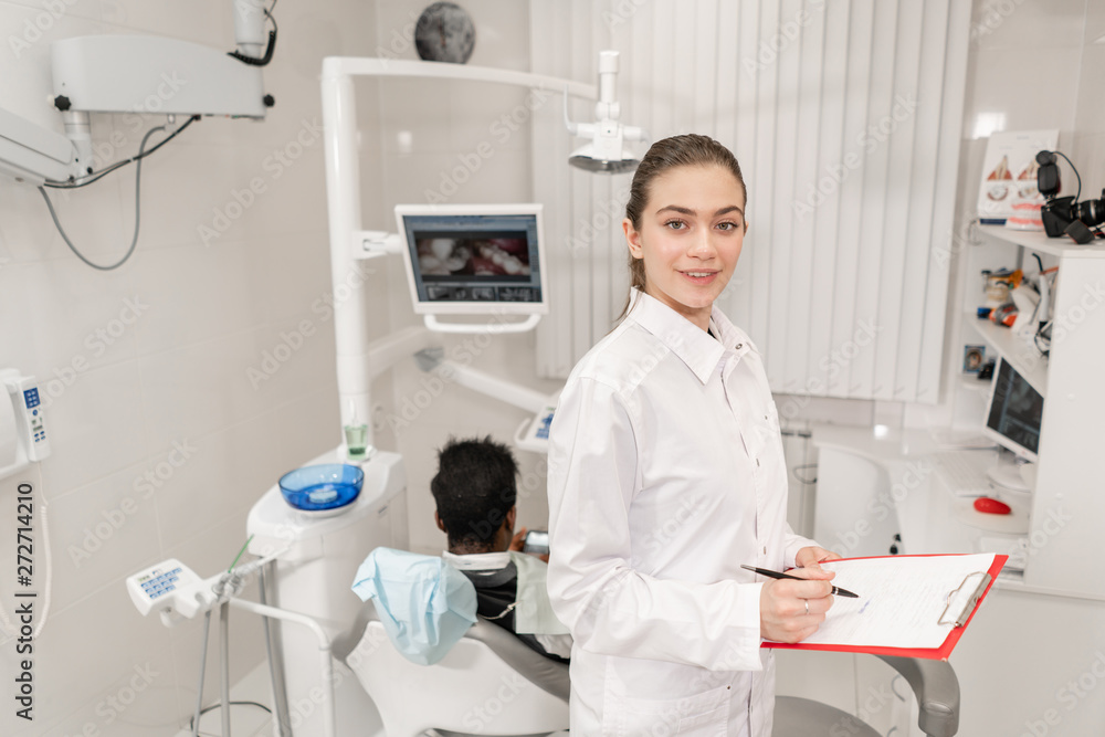 Charming beautiful girl dentist looks into the camera. Young African American male patient at chair at dental clinic. Medicine, health, stomatology concept. dentist conducts inspection and concludes. Charming beautiful girl dentist looks into the camera. Young African American male patient at chair at dental clinic. Medicine, health, stomatology concept. dentist conducts inspection and concludes.