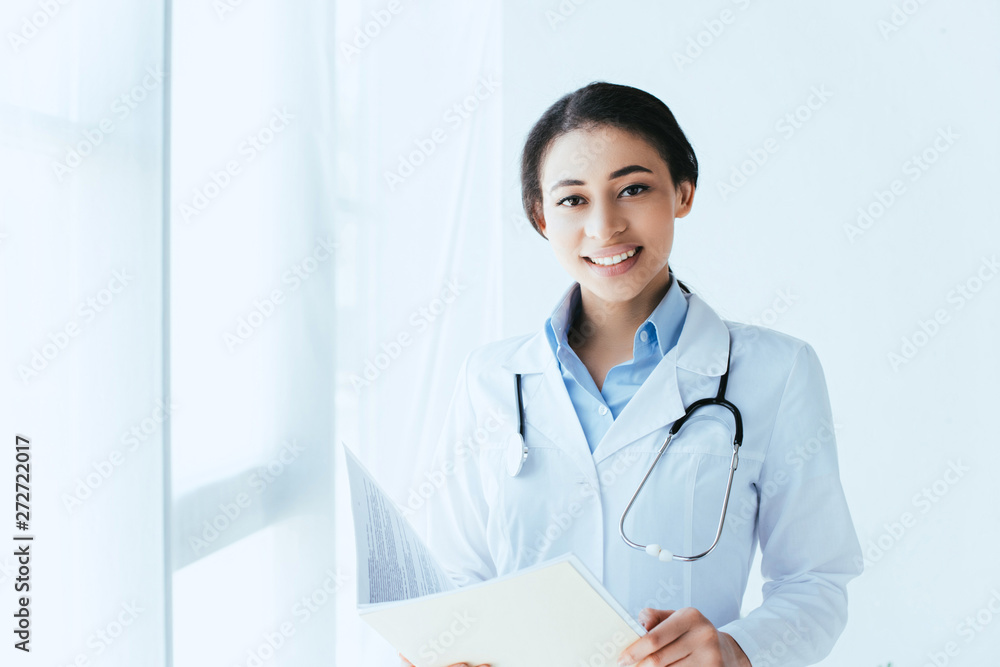 cheerful latin doctor with stethoscope holding paper folder and smiling at camera cheerful latin doctor with stethoscope holding paper folder and smiling at camera