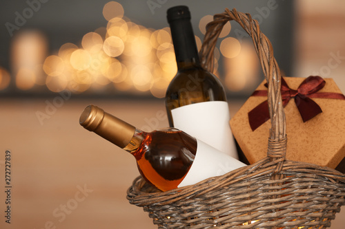 Wicker basket with bottles of wine and gift box against blurred background, closeup
