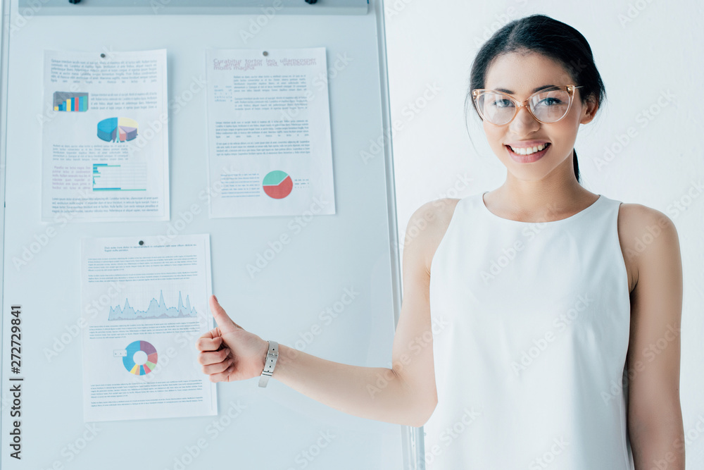 attractive businesswoman showing thumb up while standing near flipchart with infographics attractive businesswoman showing thumb up while standing near flipchart with infographics