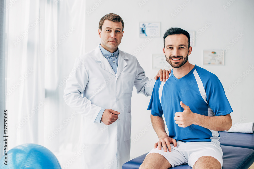 Obraz premium smiling Physiotherapist near football player showing thumb up in hospital