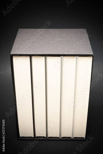 A set of five monochromatic cloth-bound books kept inside a black hard cardboard slipcase set on a slightly graduated and subtly textured dark background.