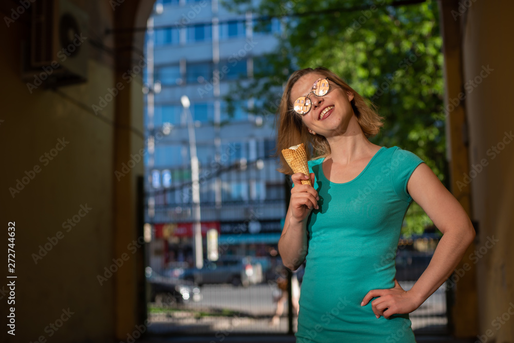 A happy woman in a turquoise dress stands in an arched passage and eats a waffle cone on a warm summer day. Beautiful blonde in sunglasses enjoys ice cream while walking. Summer dessert.