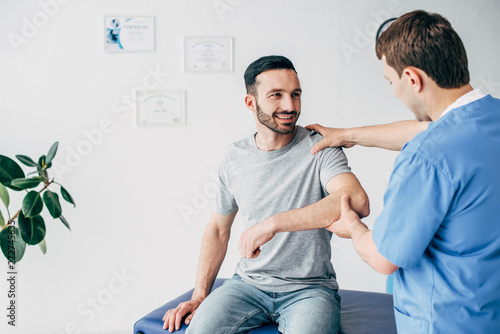Smiling patient sitting on couch and doctor examining patient shoulder in massage cabinet at clinic
