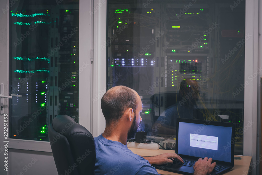 A programmer with a laptop sits in front of the glass wall of the server room. Specialist works in a modern data center. A man is sitting in a dark room with a lot of computer equipment. A programmer with a laptop sits in front of the glass wall of the server room. Specialist works in a modern data center. A man is sitting in a dark room with a lot of computer equipment.