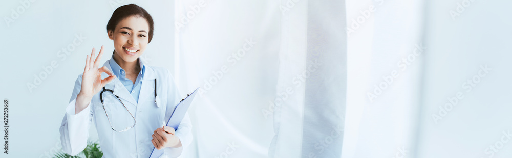 panoramic shot of happy latin doctor showing ok gesture and holding clipboard