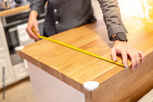 Asian man interior designer using tape measure for measuring size of wooden countertop in modern kitchen showroom in furniture store. Shopping material design for home improvement.