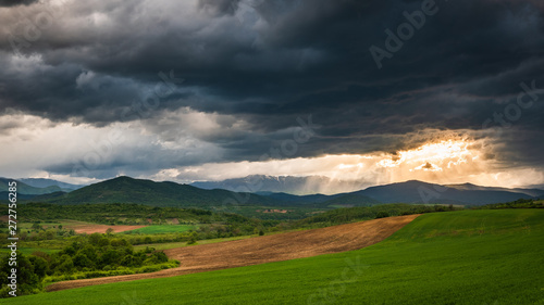A dramatic nature scenic of a green field valley with some bush and distant mountain range under a dark clouds sky at sunset
