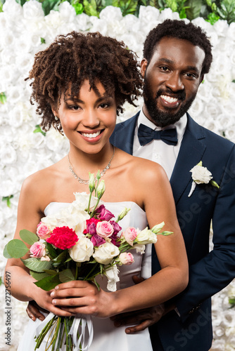 cheerful african american bride holding bouquet with flowers near bridegroom