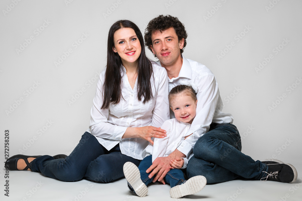Parents and their daughter are sitting together on a white background. Little girl lay down on her father. The concept of happy parenting and happy childhood.