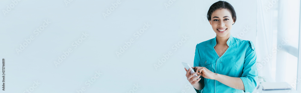 panoramic shot of young latin doctor using smartphone while smiling at camera panoramic shot of young latin doctor using smartphone while smiling at camera