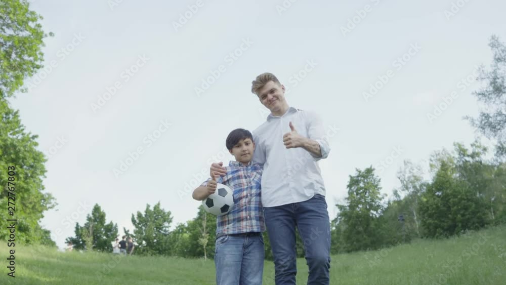 The child holding soccer ball hugging with the dad looking in camera in the park. Family leisure outdoors. The boy and his dad showing thumbs up. Father and a kid spend time together. The child holding soccer ball hugging with the dad looking in camera in the park. Family leisure outdoors. The boy and his dad showing thumbs up. Father and a kid spend time together.