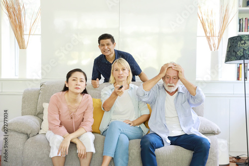 Unhappy elder multiethnic family sitting and watching television on grey sofa in living room. They expressed sadness when the football team they cheered was beaten.