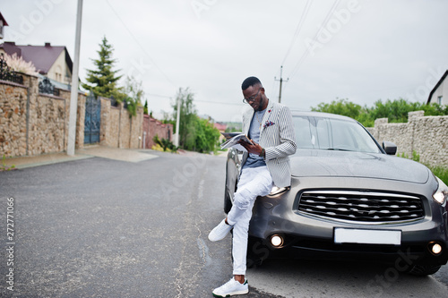 Rich and stylish african american man in blazer and white pants, eyeglasses read magazine against his suv car.
