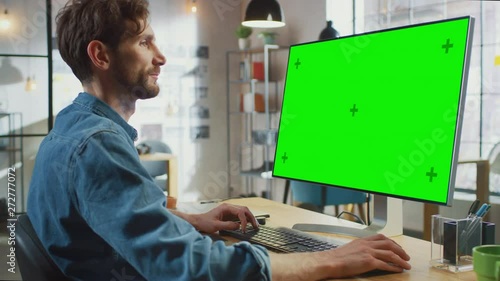 Male Creative Designer with Beard and Jeans Shirt Works on His Personal Computer with Big Green Screen Mock Up Display. He Works in a Cool Office Loft. Female Creative Colleague Walks in Background. photo