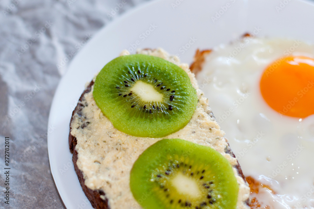 Healthy and tasty breakfast, wholemeal bread sandwich with kiwi and a fried egg on a white plate, close-up, top view Healthy and tasty breakfast, wholemeal bread sandwich with kiwi and a fried egg on a white plate, close-up, top view