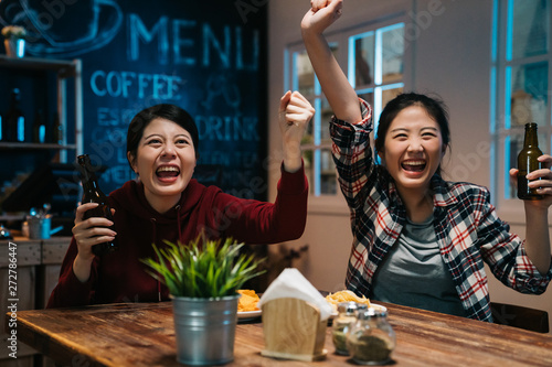 Fans watching football on TV in beer pub. excited young asian girls friends raised hands fists and screams in night sport bar. Concept of emotion experience soccer team win victory in olympic match.