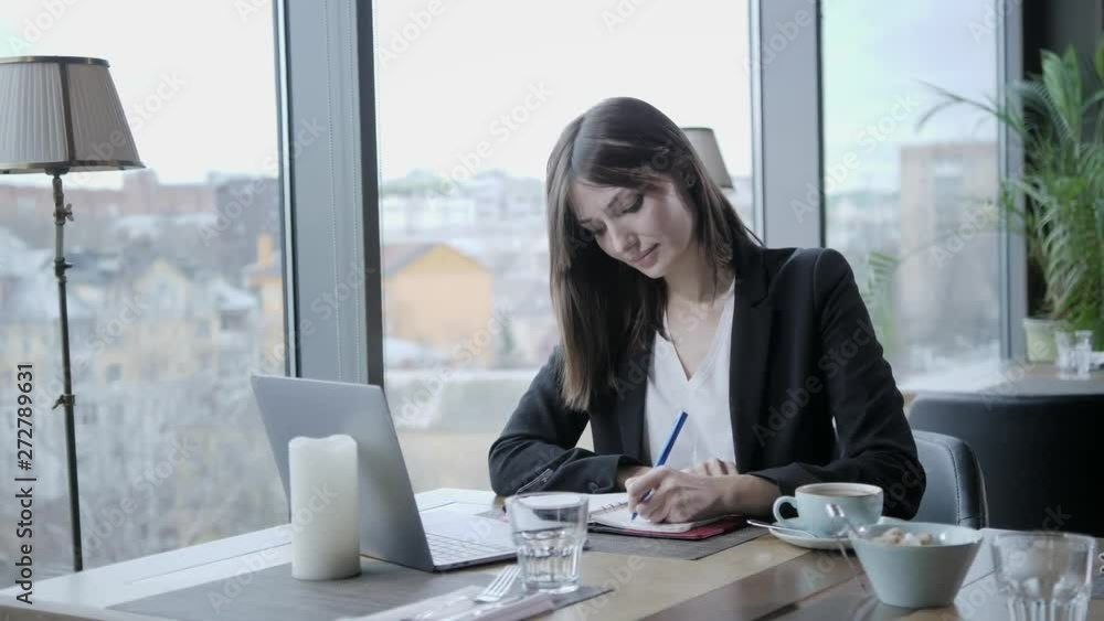 pretty woman making notes in a notebook. Young businesswoman Sitting in coffee shop at wooden table. On table is gray aluminum laptop. Schedules and makes important notes pretty woman making notes in a notebook. Young businesswoman Sitting in coffee shop at wooden table. On table is gray aluminum laptop. Schedules and makes important notes