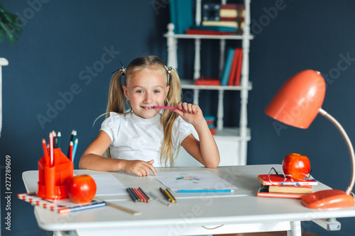 Pretty happy little girl seated at her pink desk chewing her pencil looking up at the ceiling daydreaming with a beautiful smile on her face.