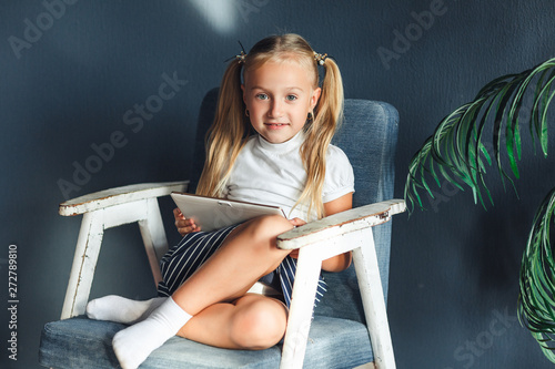 little blondy girl sitting on a chair and doing homework for school, researching information on the tablet