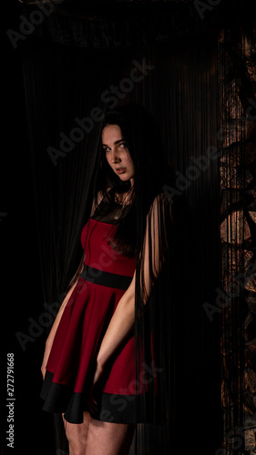 Pretty young sexy model female with dark hair in amazing long red dress and black shoes posing in dark studio