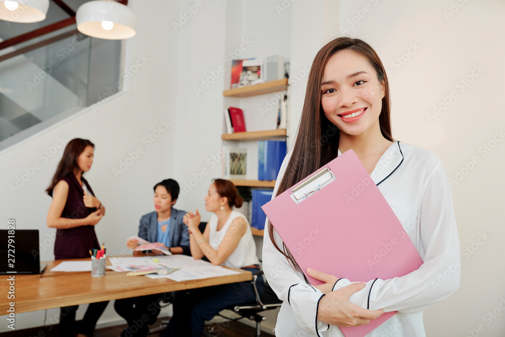 Portrait of beautiful Asian businesswoman standing with clipboard and smiling while her colleagues working in team in the background Portrait of beautiful Asian businesswoman standing with clipboard and smiling while her colleagues working in team in the background