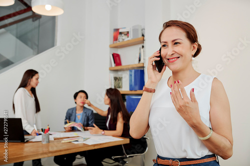 Smiling mature businesswoman standing and talking on mobile phone with her coworkers sitting at the table and discussing behind her back