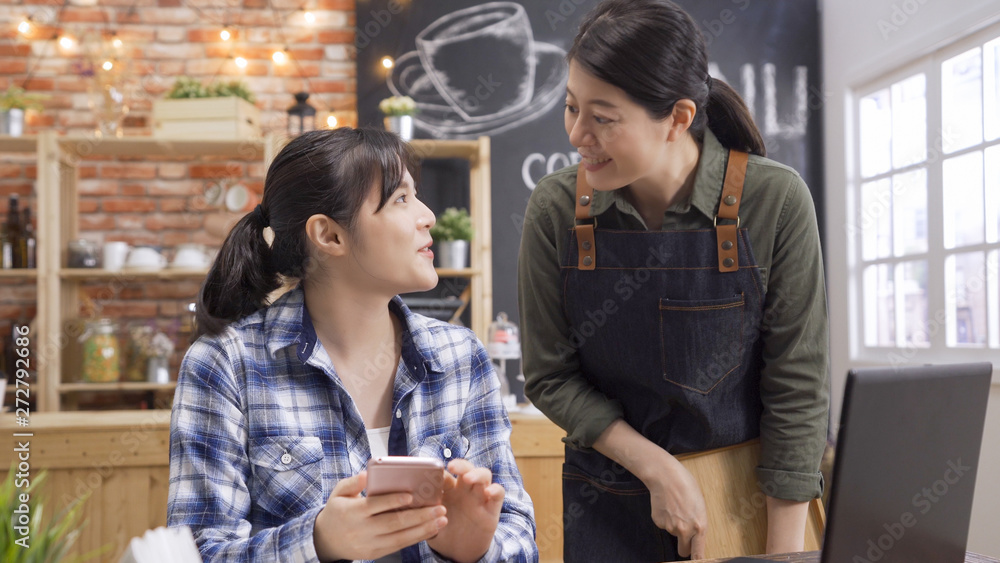 Attractive young waitress talking with regular customer friend. girl client using mobile phone chatting with barista while take order in coffee shop. two smiling women friendship having conversation. Attractive young waitress talking with regular customer friend. girl client using mobile phone chatting with barista while take order in coffee shop. two smiling women friendship having conversation.