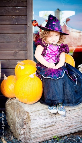 Happy Halloween! Cute cheerful little witch with a magic wand. Beautiful child girl in witch costume sitting on the big pumpkin, conjuring and laughing.