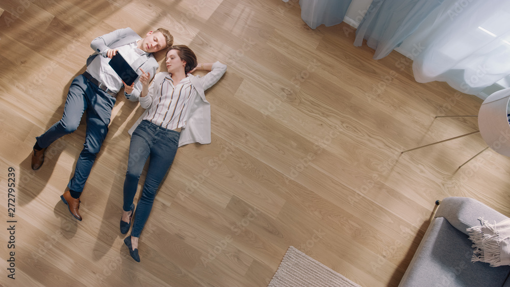Young Couple are Lying on a Wooden Flooring in an Apartment. They are Using a Tablet Computer. Cozy Living Room with Modern Interior, Grey Sofa and Wooden Parquet. Top View Camera Shot. - obrazy, fototapety, plakaty 