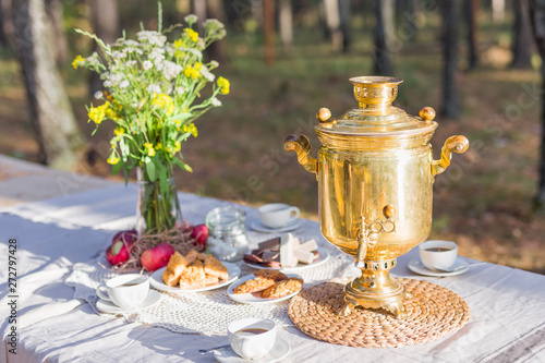 Golden russian samovar with tea cups, snacks on table decorated with some flowers outdoors