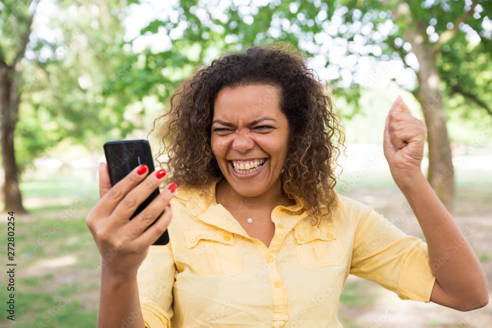 Cheerful woman using smartphone in city park. Young lady pumping fist and standing with blurred green trees and lawn in background. Win concept. Front view. Cheerful woman using smartphone in city park. Young lady pumping fist and standing with blurred green trees and lawn in background. Win concept. Front view.