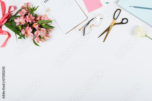 top view of flowers with ribbon, clipboard, scissors, notebook, pencil and glasses on white
