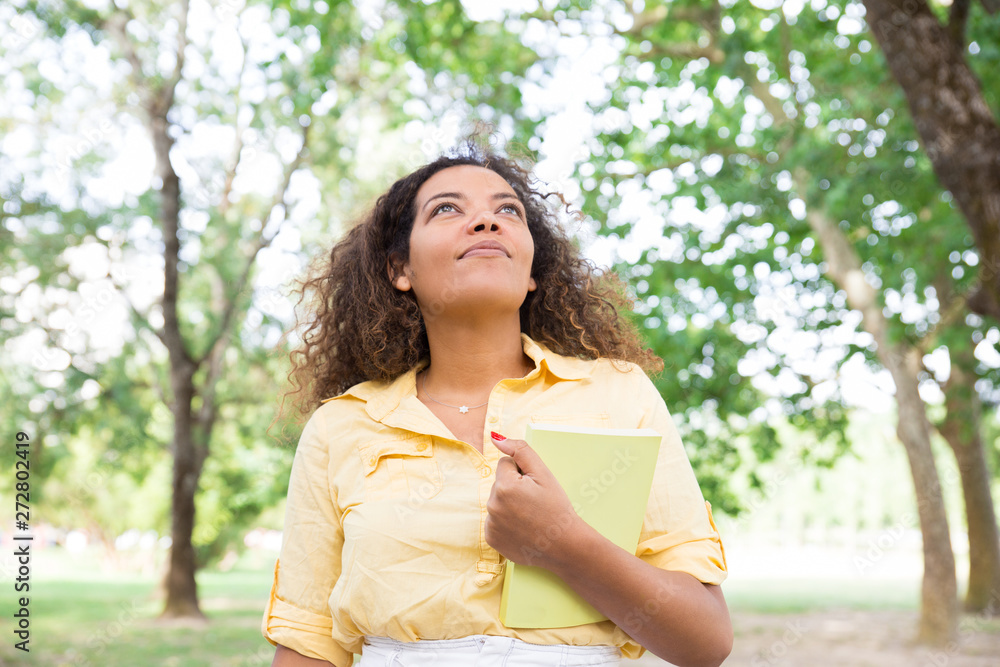 Dreamy young woman holding book in city park. Woman looking upwards and standing with blurred trees in background. Summer and education concept. Front view. Dreamy young woman holding book in city park. Woman looking upwards and standing with blurred trees in background. Summer and education concept. Front view.